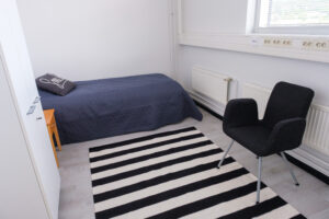 Asuntolahuone, jossa sänky, vaatekaappi, raidallinen matto ja nojatuoli