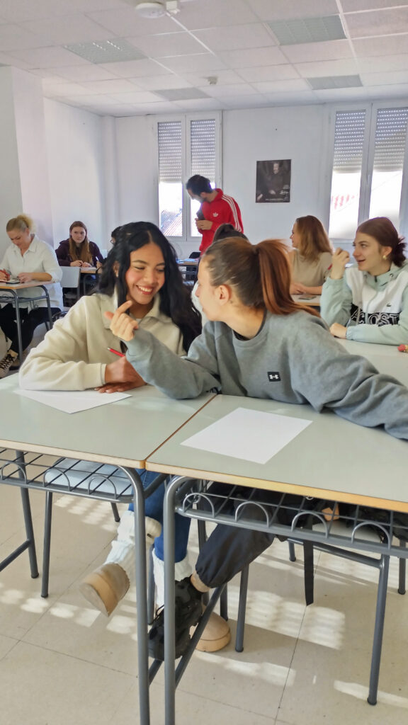 Etualalla kaksi nuorta oppitunnilla, vaihtavat nauraen ajatuksia. Taustalla opettaja ja muita aikuisopiskelijoita.