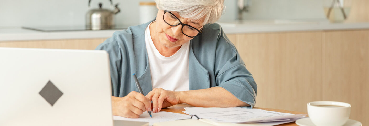 Aikuinen kypsässä iässä kirjoittaa kotinsa pöydän ääressä, pöydällä tietokone ja muistikirja